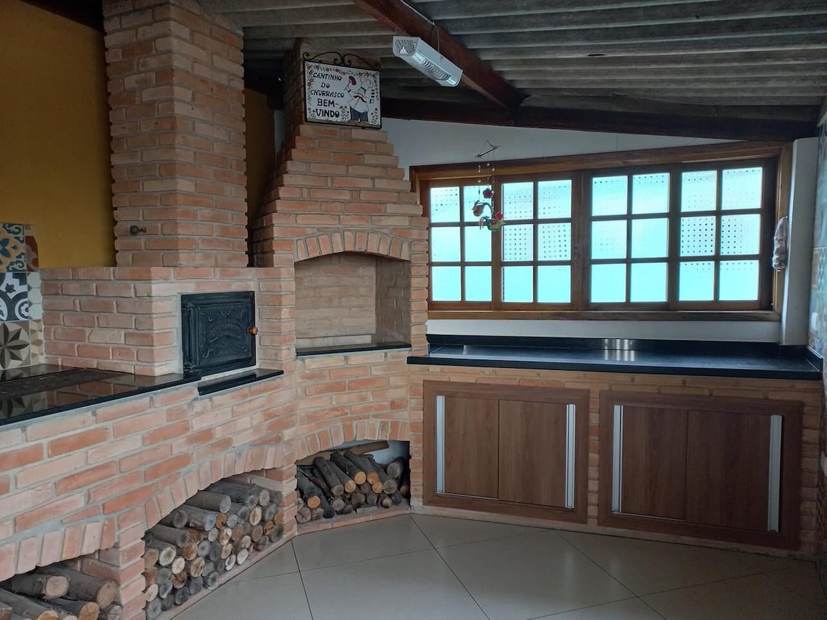 A spacious kitchen features a traditional brick barbecue area complemented by a wood-burning stove. Ample counter space is provided alongside wooden cabinetry. Natural light filters through multiple small window panes, illuminating the area while showcasing neatly stacked firewood below the barbecue.