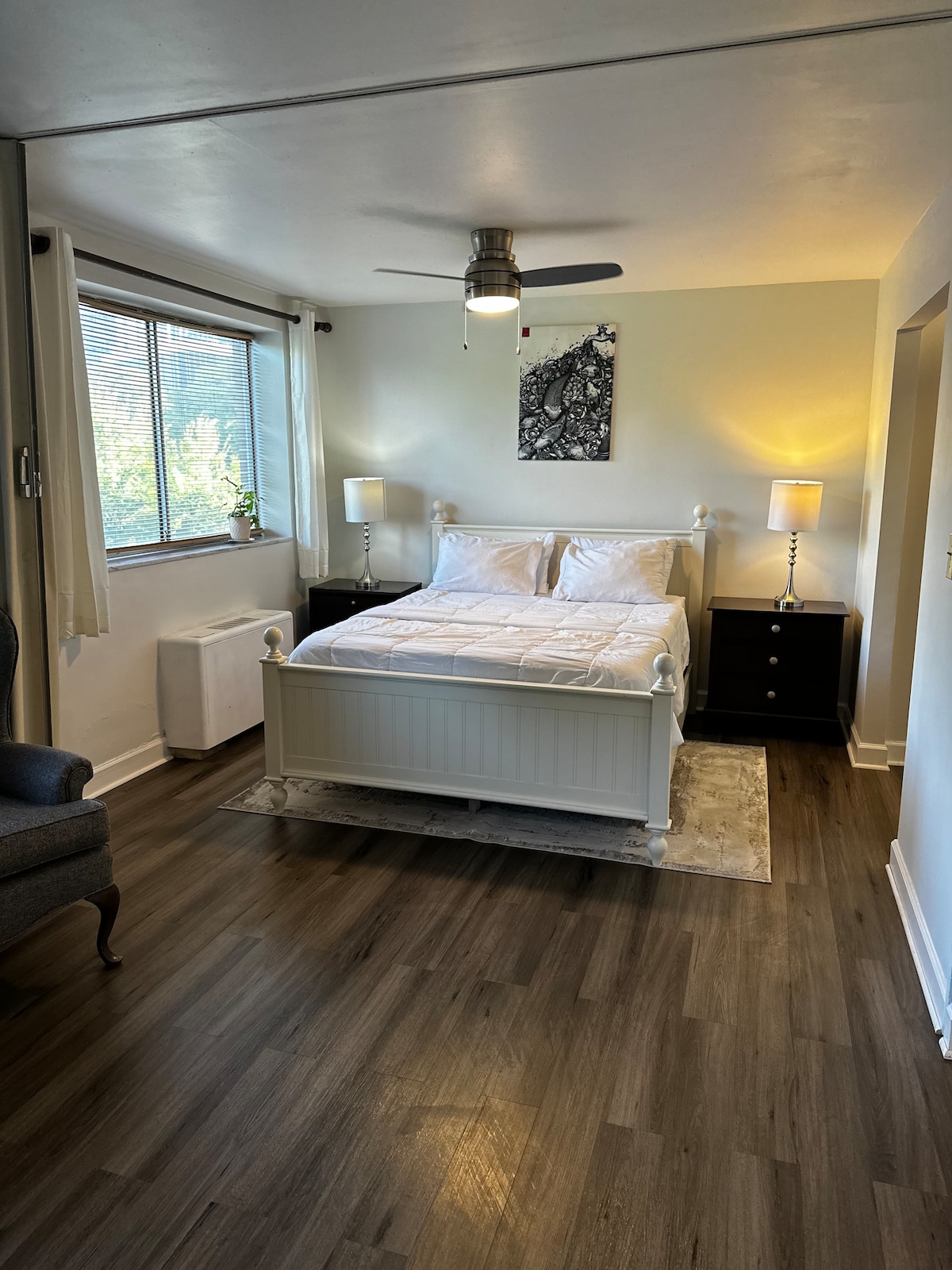 A well-lit bedroom features a spacious bed with crisp white linens centered against a window. Two bedside tables with lamps flank the bed. A comfortable chair is positioned nearby, and a piece of artwork adorns the wall above the bed, adding visual interest to the space.