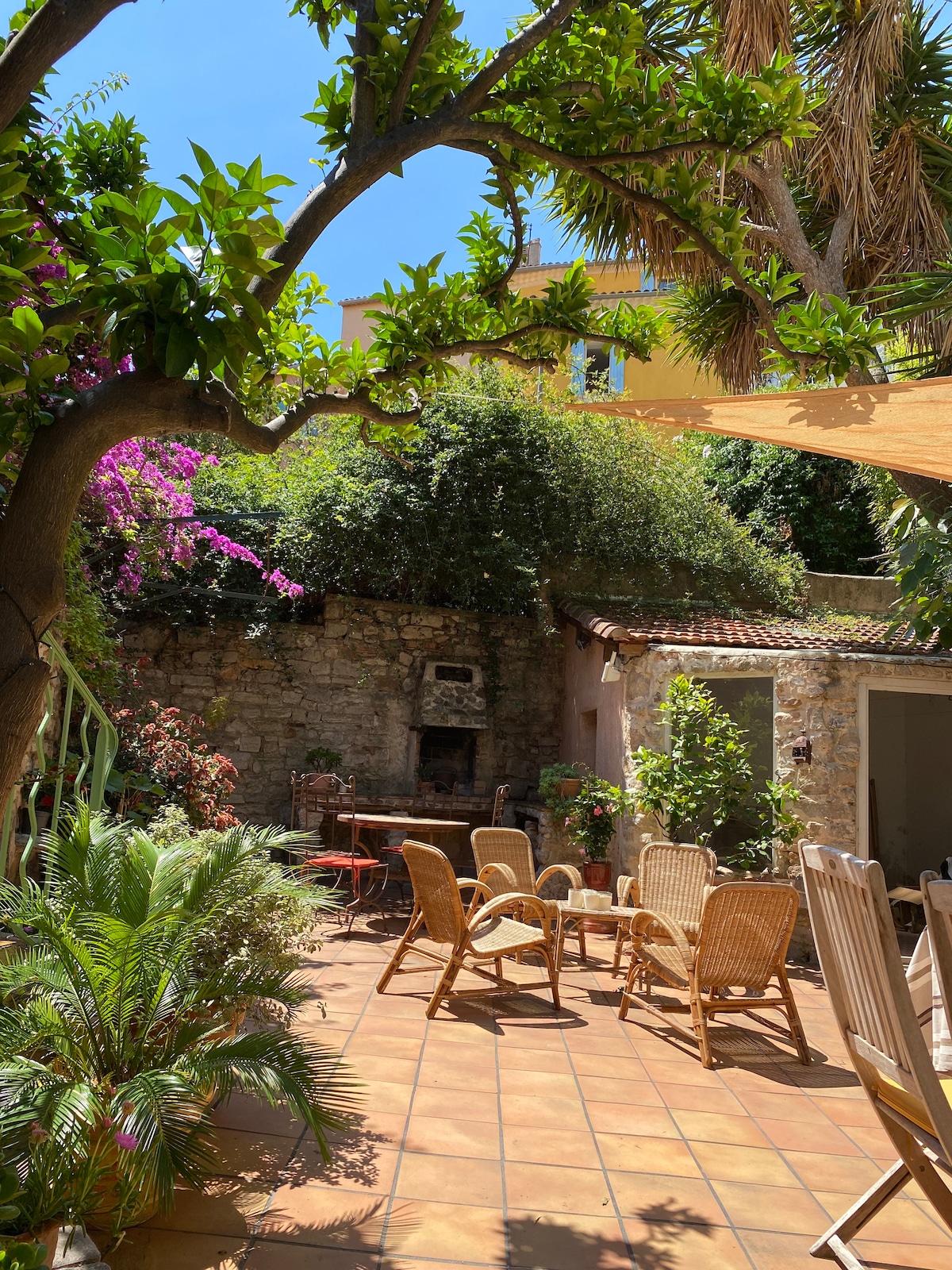 A serene courtyard is surrounded by vibrant greenery, including a large tree and blooming flowers. A cozy seating area features several wicker chairs and a small table, set on terracotta tiles. A rustic stone wall provides privacy, with a barbecue area and storage space visible in the background.