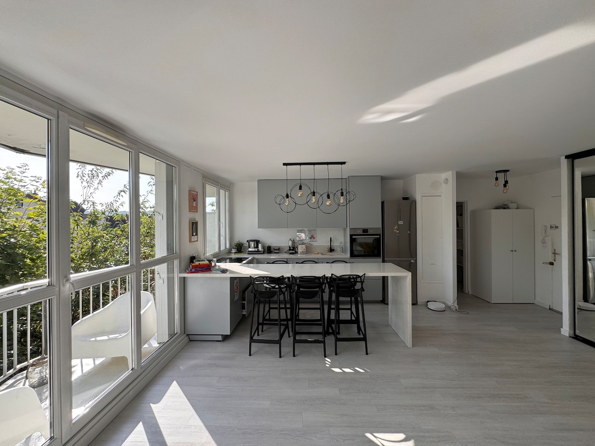 A modern kitchen area features a central island with seating for six. Large windows provide natural light and views of greenery outside. Stainless steel appliances are included, with a minimalist design and ample storage space. A doorway leads toward additional living areas.