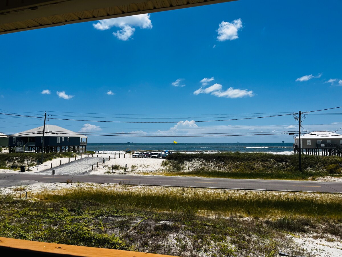 A view of the sparkling Gulf of Mexico is visible from the porch, framed by grassy dunes and white sandy beach. Clear blue skies stretch overhead, dotted with a few clouds. Nearby structures and a road are seen, enhancing the coastal ambiance.