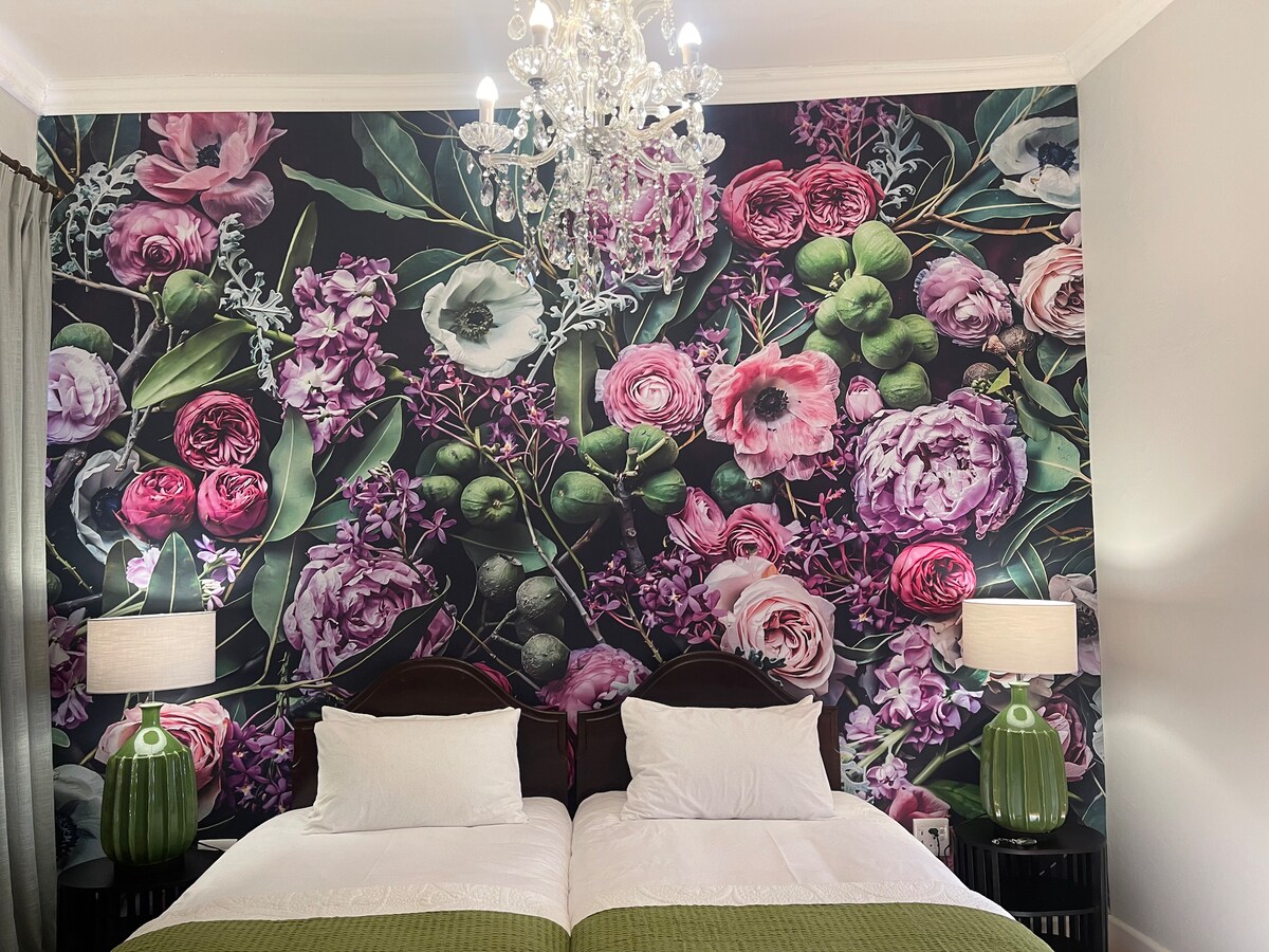 A vibrant floral wallpaper adorns the wall behind two single beds, each dressed in white linen. Decorative green lamps sit on dark nightstands beside the beds, and a chandelier hangs from the ceiling, providing soft ambient lighting.