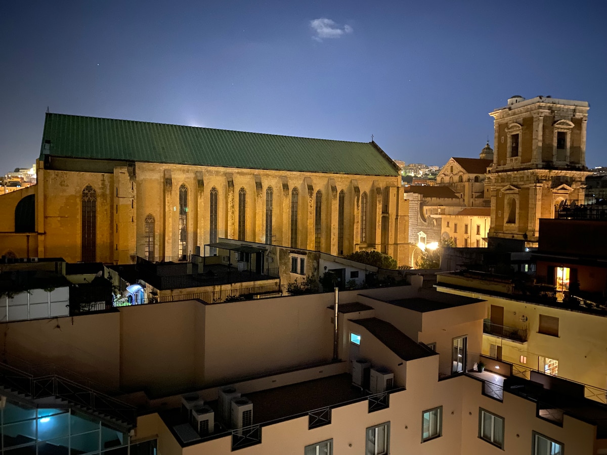 The image captures a nighttime view of historic architecture, featuring a large building with a green-roofed structure and prominent stained glass windows. Nearby, a well-lit smaller building with an ornate facade adds to the urban landscape, against a backdrop of a starry sky.