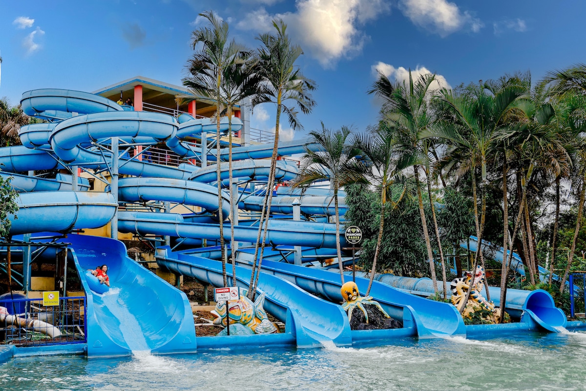 A collection of twisting blue water slides forms part of a vibrant water park. Lush palm trees provide a tropical ambiance as water splashes from the slides into a pool below. A multi-story structure is visible in the background, adding to the park's recreational offerings.