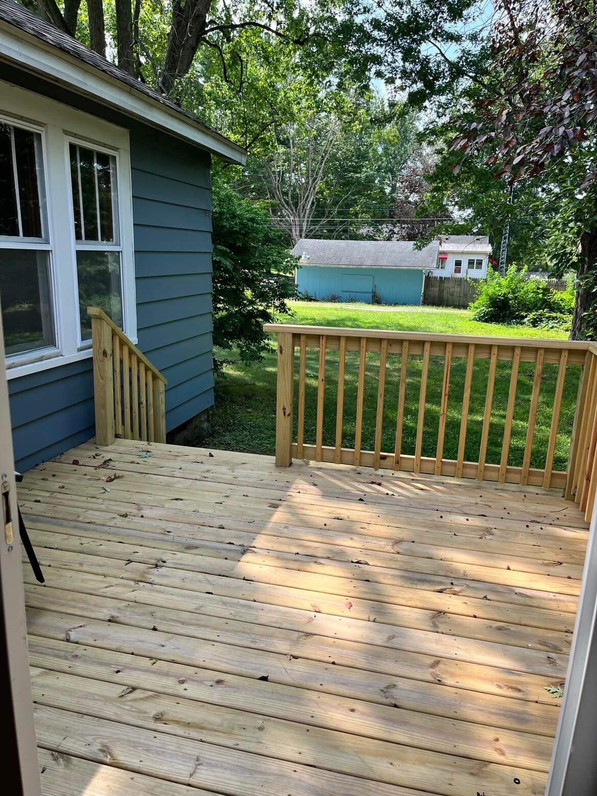 A wooden deck is shown, featuring a railing and overlooking a grassy area. Sunlight is visible on the surface, indicating a bright day. Lush greenery can be seen in the background, adding to the natural setting.