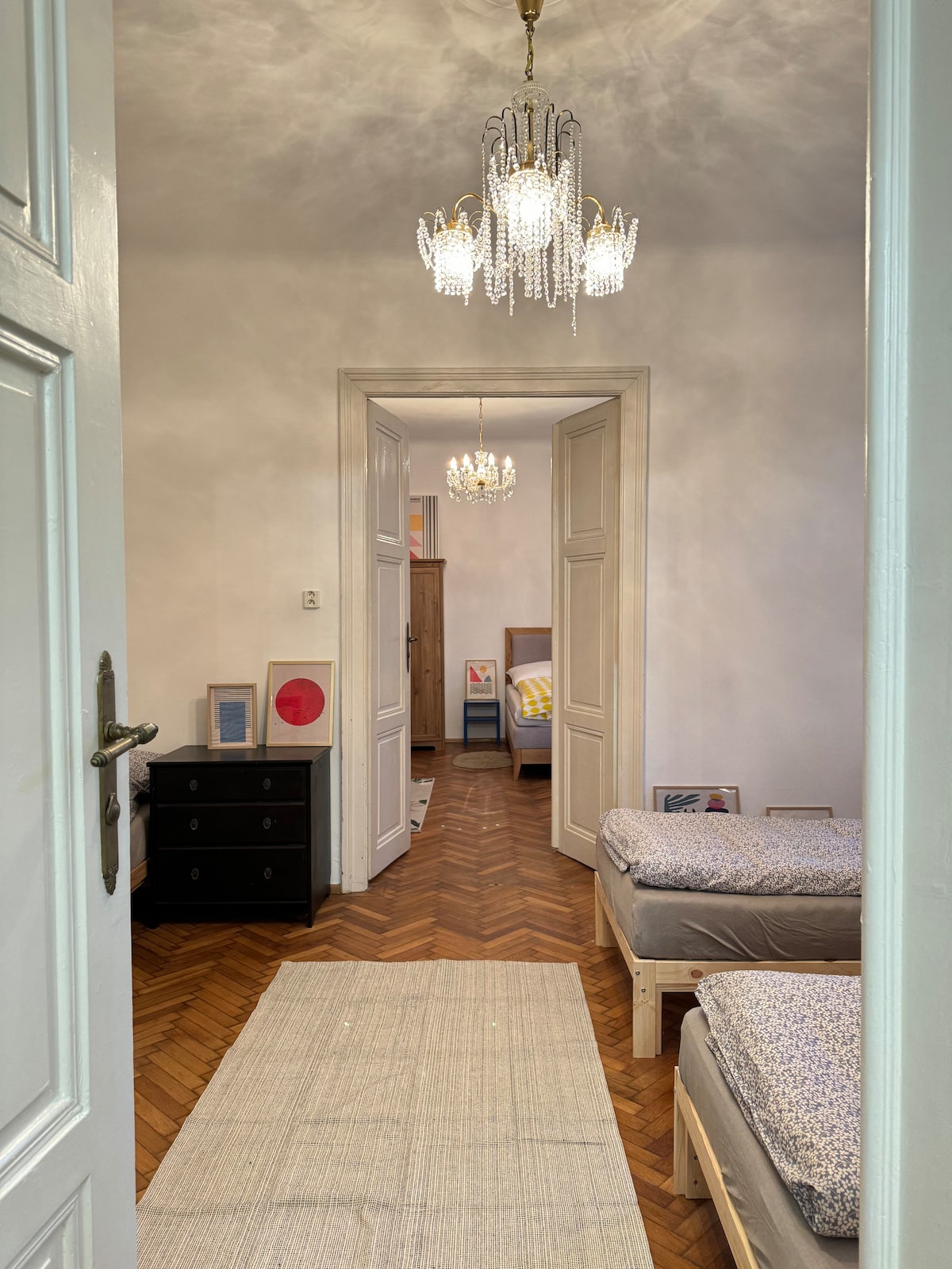 An inviting hallway leads to two bedrooms, each furnished with separate beds. Elegant chandeliers hang from the ceiling, illuminating the space. A light rug lines the wooden floor, contributing to a calm atmosphere while doorways offer glimpses into the adjacent rooms.