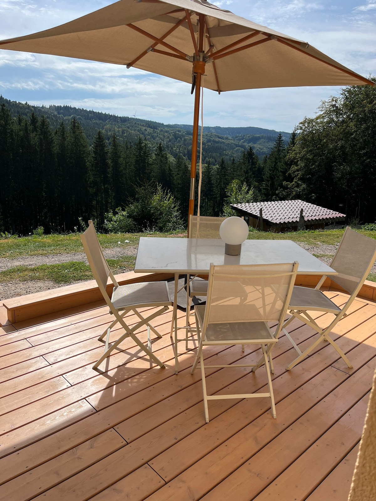 An outdoor deck features a table surrounded by four chairs, shaded by a large umbrella. A soft light source is positioned at the center of the table. The scenic backdrop showcases a lush forested hillside under a clear sky.