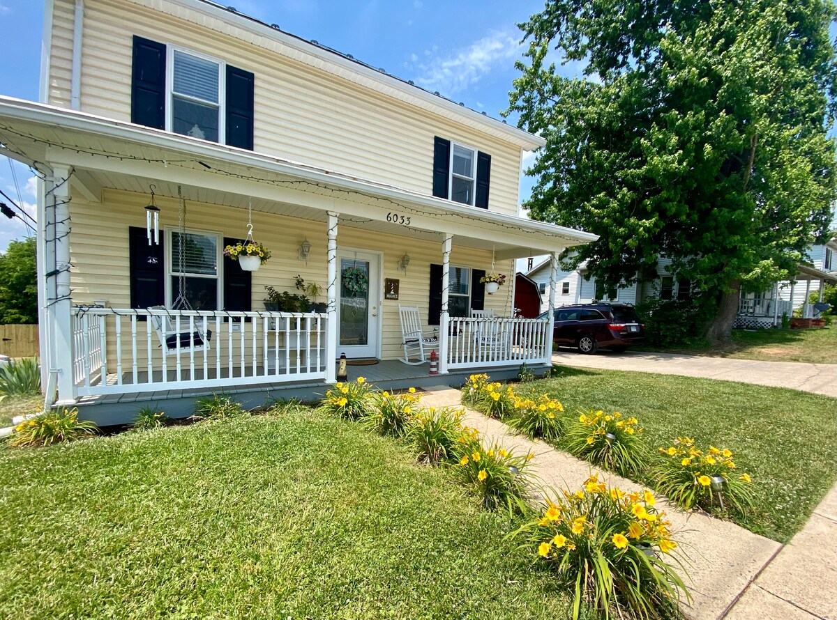 A two-story house features a welcoming front porch with white railings and rocking chairs. Lush green grass surrounds the home, accented by bright yellow flowers. Large trees provide shade, and a car is parked in the driveway. The house displays a light-coloured exterior with dark shutters.
