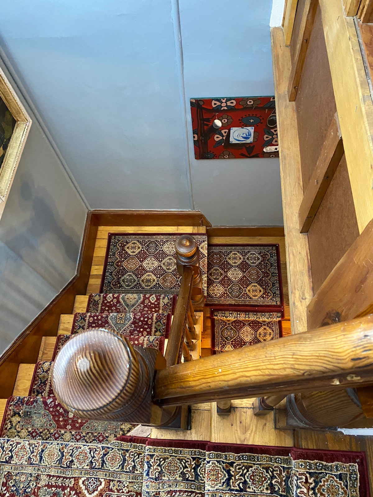A wooden staircase leads down from the second floor, bordered by intricately patterned rugs that add warmth to the setting. The bannister features rounded wooden posts, and an ornate piece of wall art is visible near the ceiling.