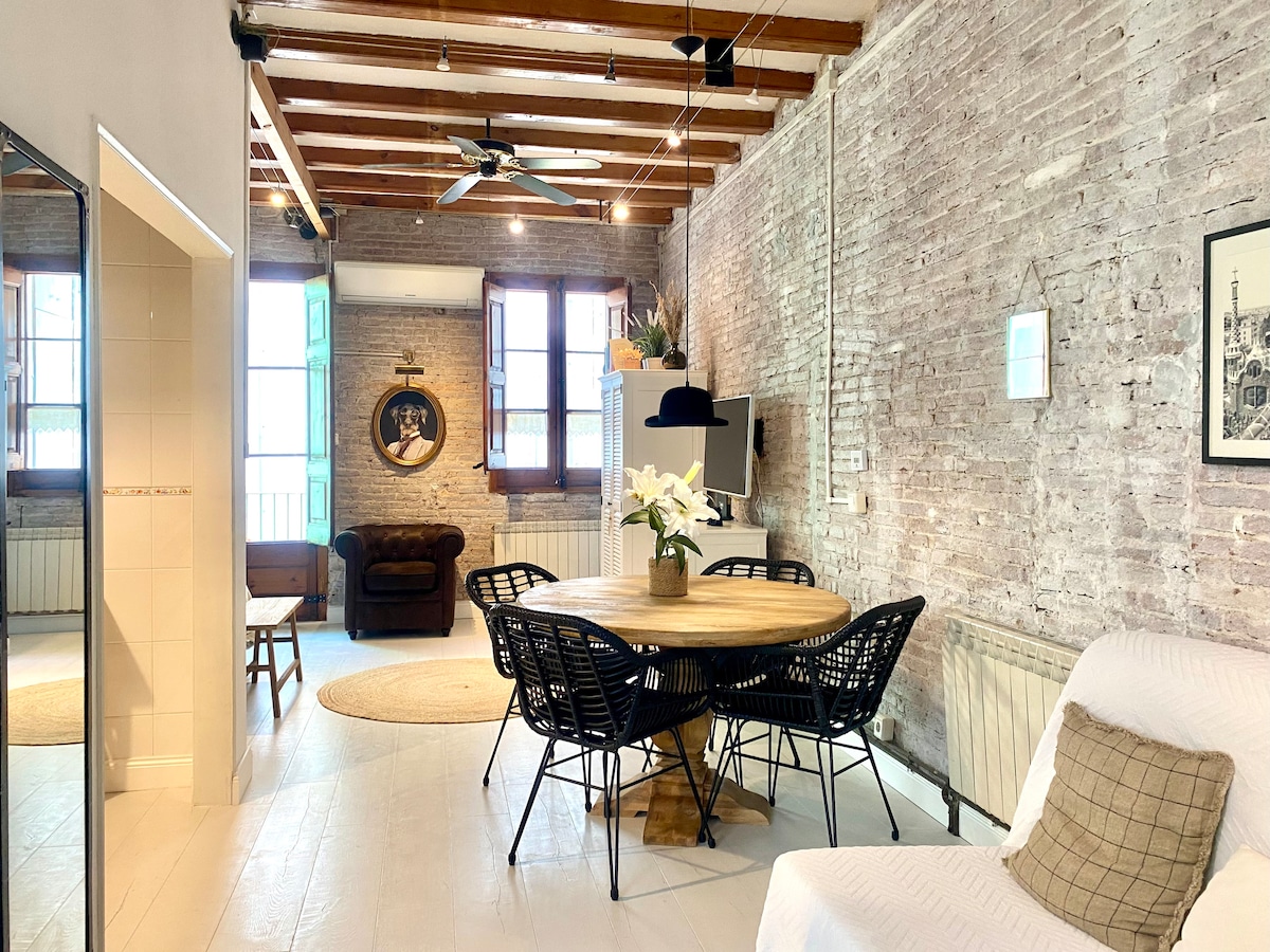 The spacious interior features exposed brick walls and wooden beams. A round dining table surrounded by black wicker chairs is centrally positioned. A comfortable seating area with a brown couch is visible, along with a decorative rug on the floor. Light streams in from large windows.