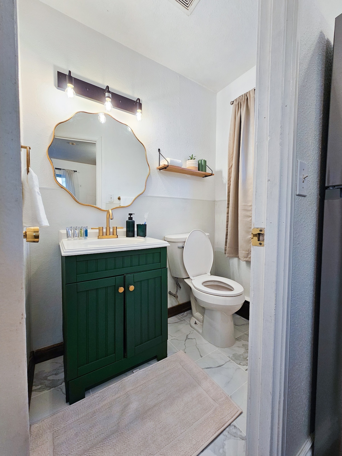 A bathroom features a green vanity with a white countertop, complemented by a round mirror above. Soft lighting illuminates the space, while beige curtains frame the window. A neutral rug lies on the marble floor, and toiletries are neatly arranged on a wall shelf.