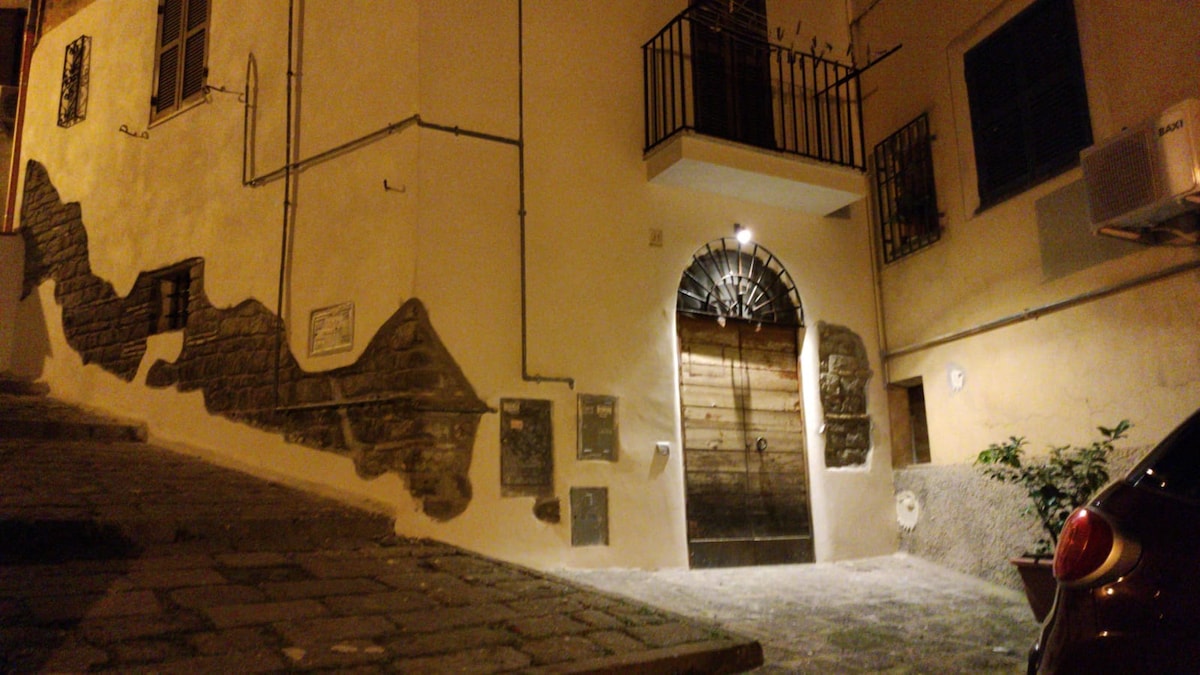 The exterior of a charming building is captured at night, featuring a wooden door with decorative wrought iron accents. The stone walls are complemented by ambient lighting, and a small plant can be seen near the entrance. Narrow cobblestone streets lead up to the building.