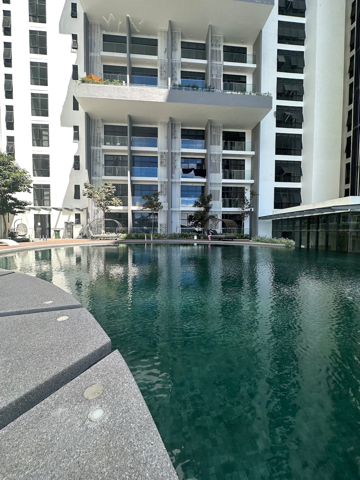 A sleek, modern building is depicted beside a clear swimming pool, surrounded by minimal landscaping. Large windows reflect natural light while balconies offer green plants. The pool's edge features smooth stone seating, creating a serene space for relaxation.