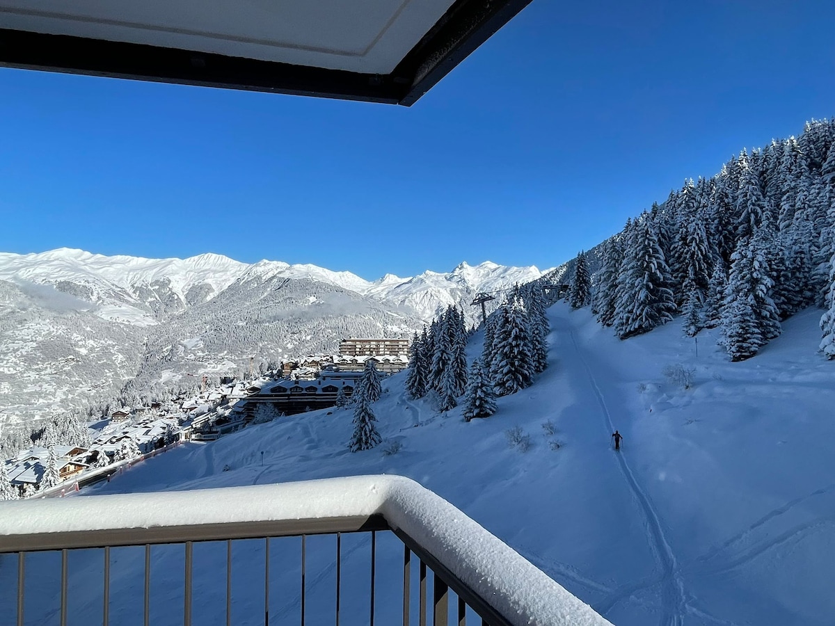 A stunning winter landscape is presented from the balcony, featuring snow-covered mountains in the distance. The foreground includes a slope marked by ski tracks, while lush evergreens blanket the nearby hills. Clear blue skies enhance the serene atmosphere of the snow-blanketed scene.