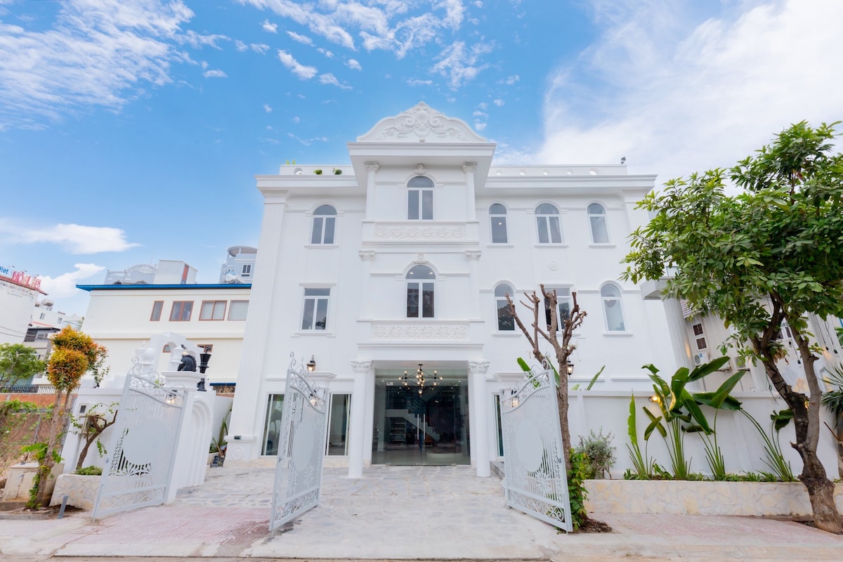 The villa's exterior features a grand white facade with ornate architectural details. A spacious entrance is framed by manicured gardens and trees, complemented by a wide pathway leading to the entrance. The sky showcases a bright blue hue with scattered clouds.