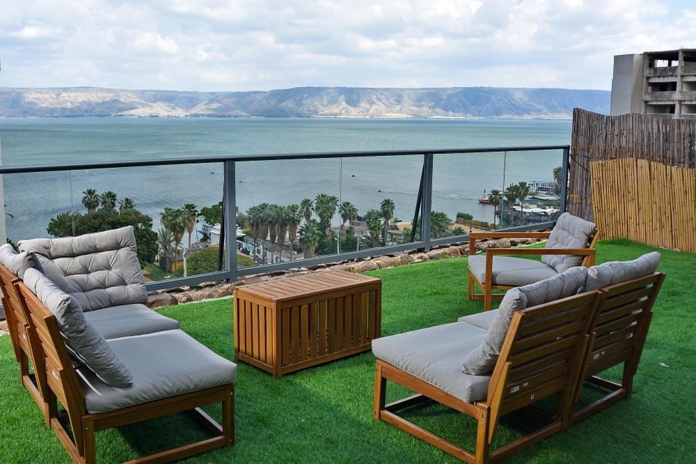 A spacious outdoor seating area is presented, featuring four comfortable lounge chairs arranged around a wooden coffee table. A lush green lawn enhances the space, while panoramic views of the Sea of Galilee and surrounding landscape are visible in the background.