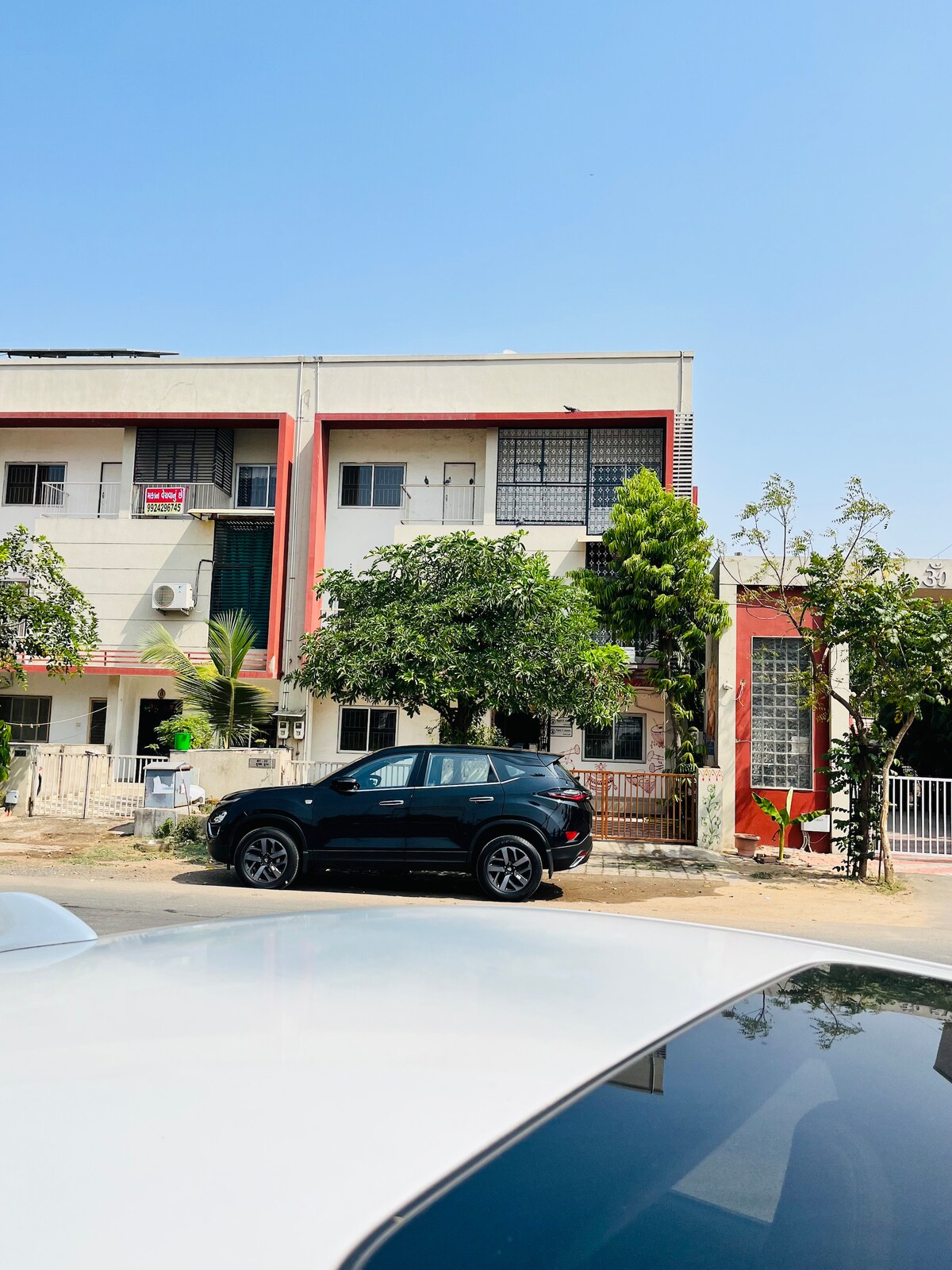 A modern multi-story building with a vibrant red accent is visible against a clear blue sky. The façade features a combination of large windows and balconies, while green trees provide a natural touch to the surrounding area. A parked black SUV is positioned in the foreground.