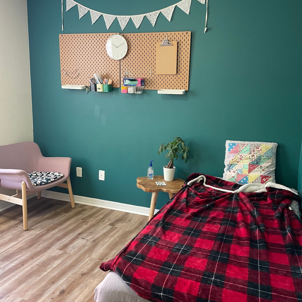A cozy room features a spacious bed with a red and black plaid blanket draped over it. A small table holds a plant, and a pink armchair is positioned nearby. A wall board displays organizational items and a clock, while the walls are painted in a calming green.