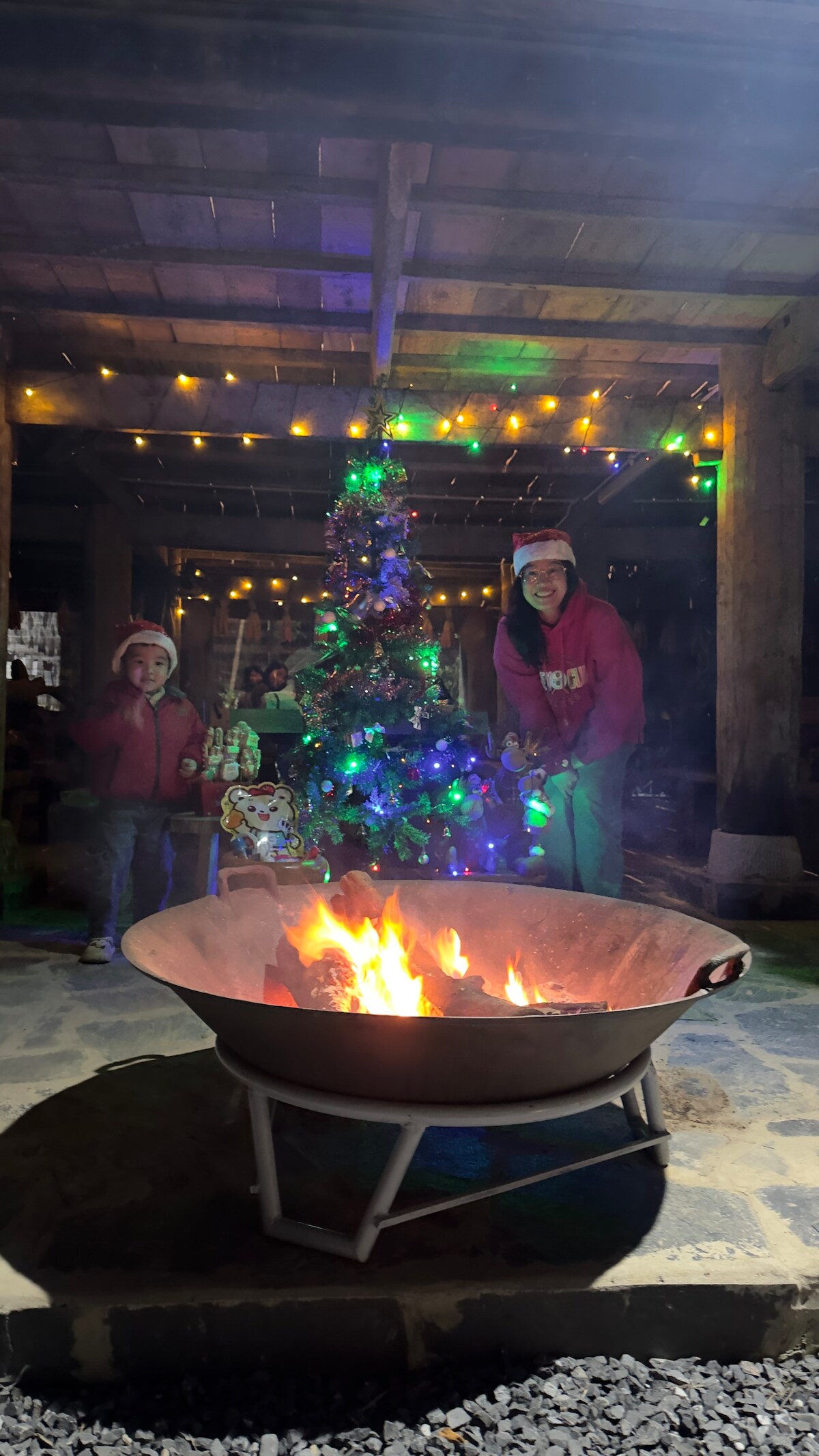 A fire pit is centered in the foreground, flames flickering warmly. A decorated Christmas tree with colorful lights stands nearby. String lights illuminate the covered outdoor area, creating a festive ambiance. Two individuals, wearing matching red jackets, enjoy the cozy atmosphere.