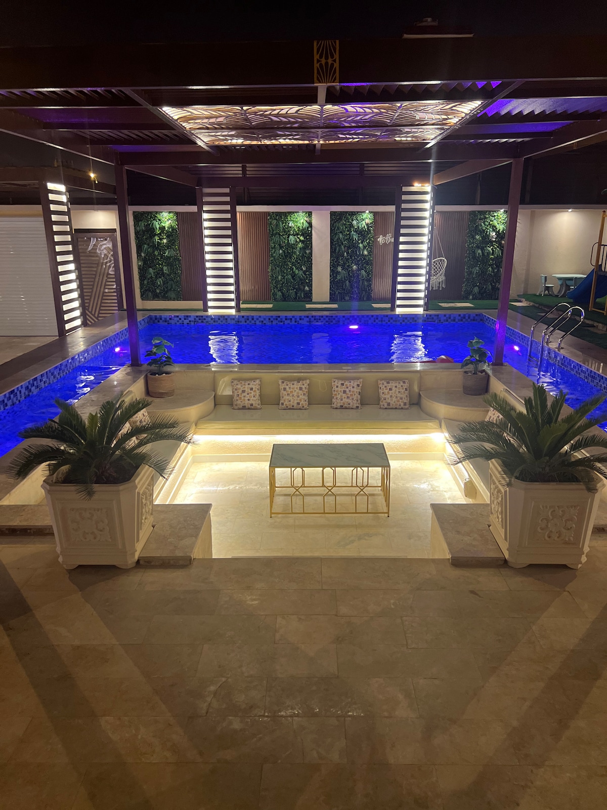 An illuminated pool area features a spacious seating arrangement with several cushions and plants. The pool itself is surrounded by elegant stone tiles. Vertical greenery is visible in the background, while soft lighting enhances the tranquil ambiance of the space.