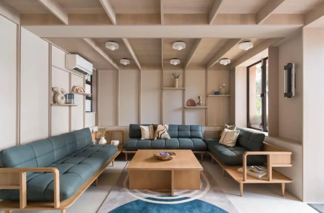 A spacious living area features a comfortable arrangement of teal sofas and a wooden coffee table at the center. Shelving units are built into the walls, displaying decorative items. Natural light is allowed in through large windows, enhancing the room's inviting ambiance.