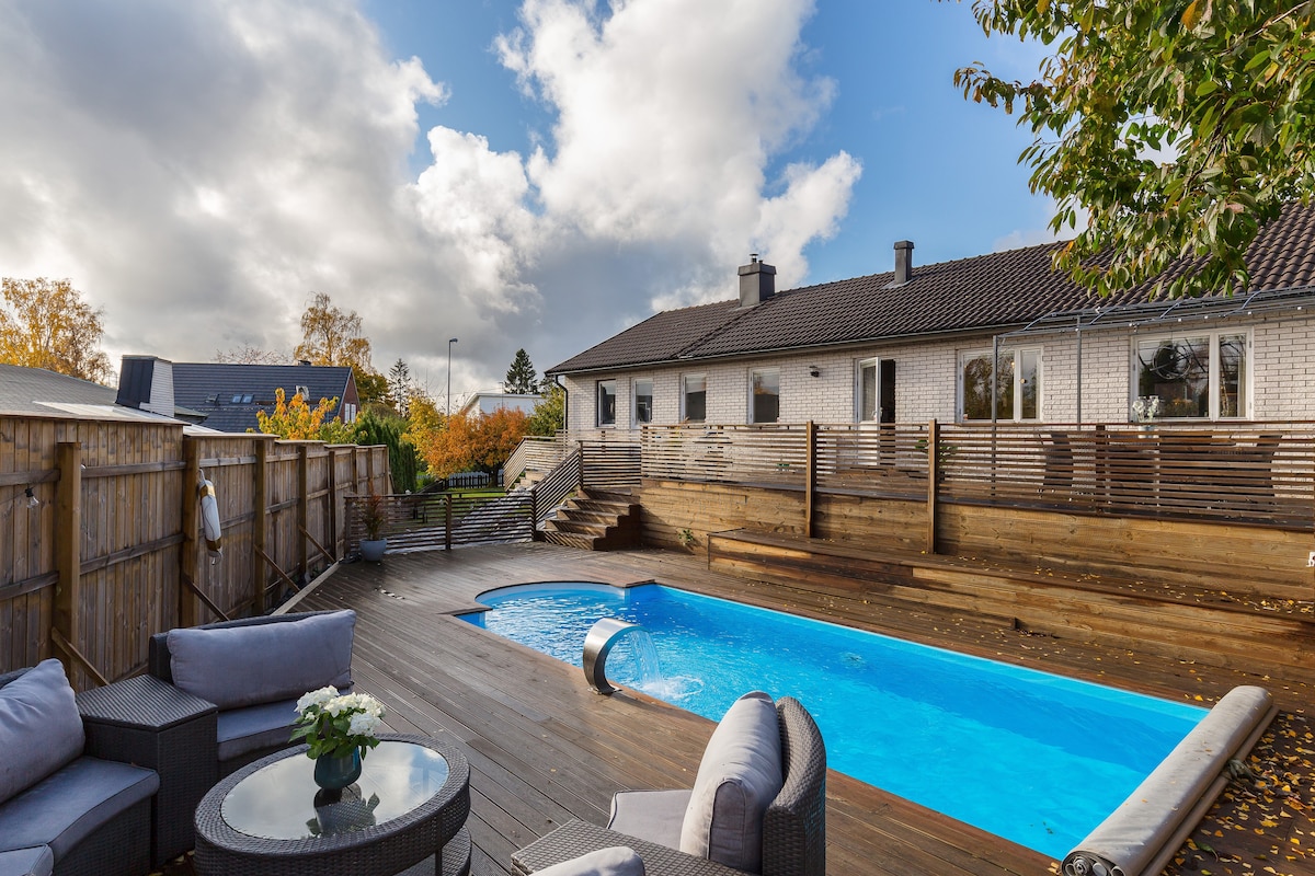 A wooden deck features a rectangular pool surrounded by lounge seating, offering a comfortable space for relaxation. Steps lead up to the adjacent building, with a mix of trees and shrubs enhancing the outdoor area. The sky is partly cloudy, creating a dynamic backdrop.