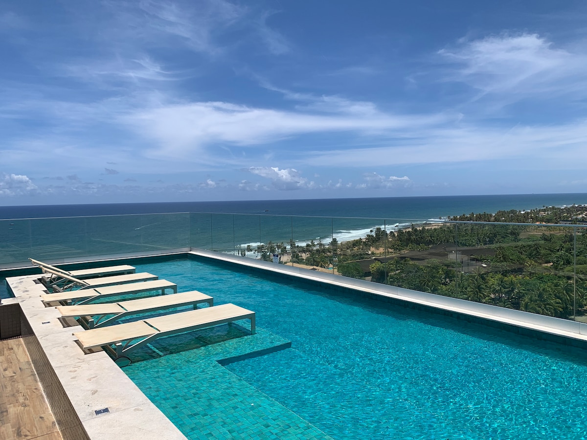 A rooftop area features a sparkling swimming pool with clear turquoise water. Sun loungers are arranged along the pool's edge, providing a serene view of the expansive ocean and coastline under a bright blue sky.