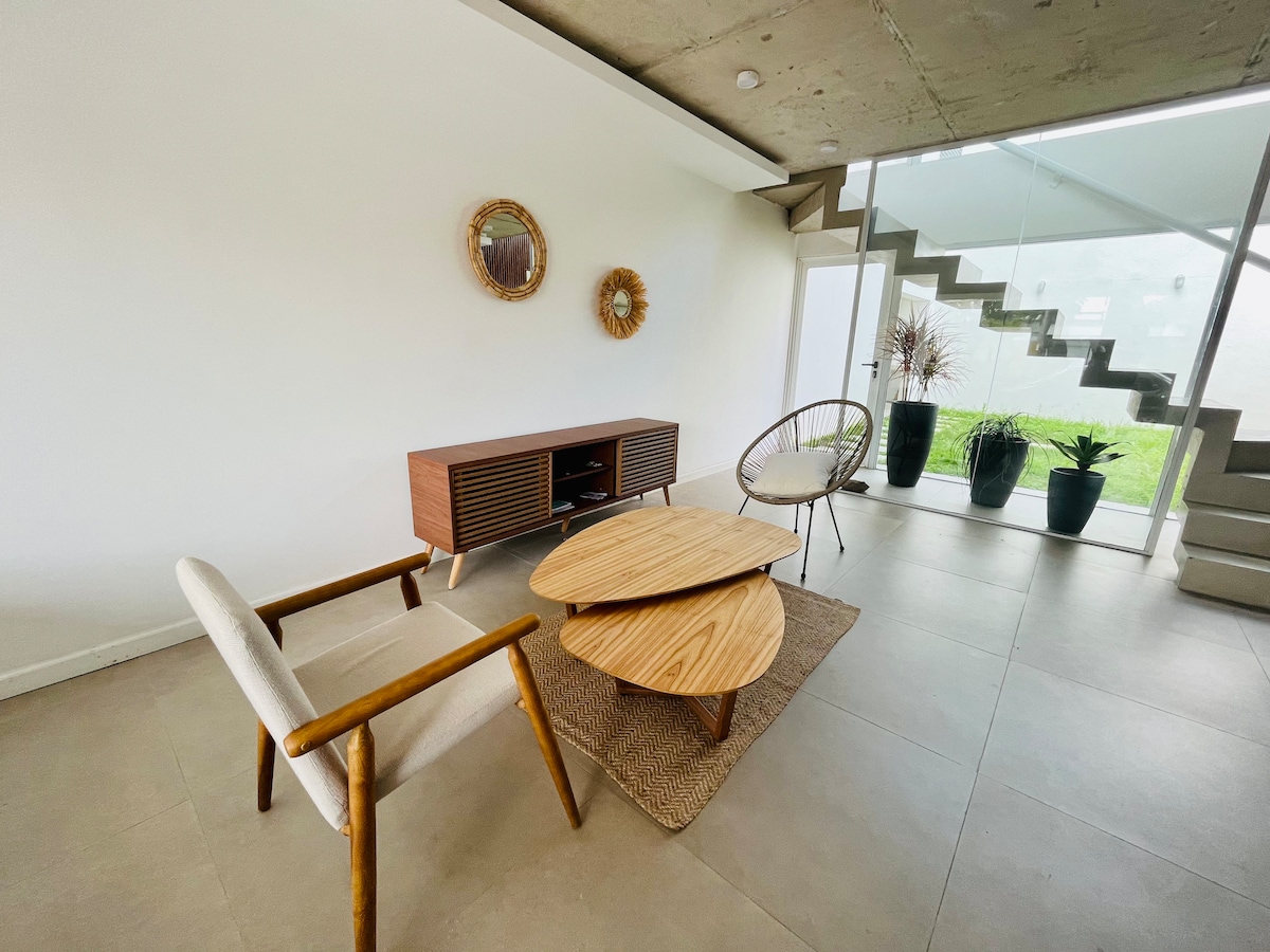 A modern living area features two stylish chairs, a round wood coffee table, and a decorative rug. The space is brightened by natural light filtering through the glass doors. Potted plants are positioned near the staircase, enhancing the green view from the interior.