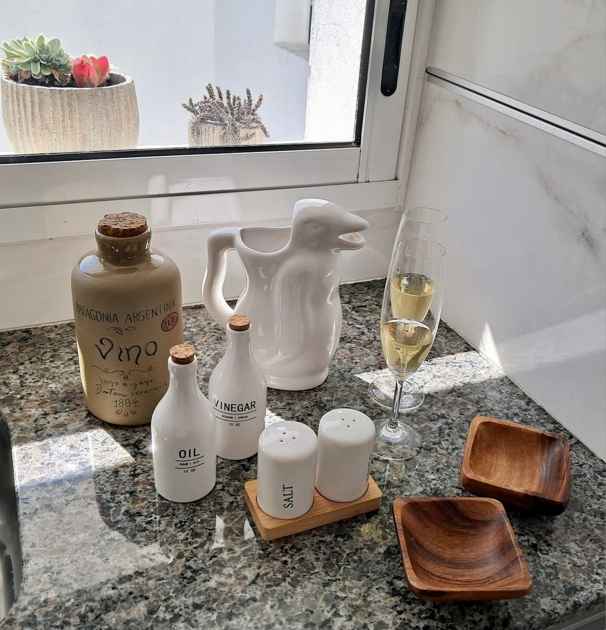 An elegant display on a kitchen counter features a light-colored pitcher and two champagne flutes. A decorative ceramic bottle labeled 'Patagonia Argentina' complements the setting, alongside white oil and vinegar bottles. Salt and pepper shakers rest on a wooden tray with two small wooden bowls.