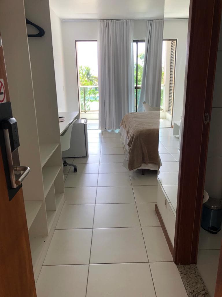 An open doorway leads into a bright room featuring a spacious bed covered with a light blanket. A large window with sheer white curtains allows natural light to fill the space, while a small desk is positioned nearby. Neutral tile flooring enhances the room's airy feel.