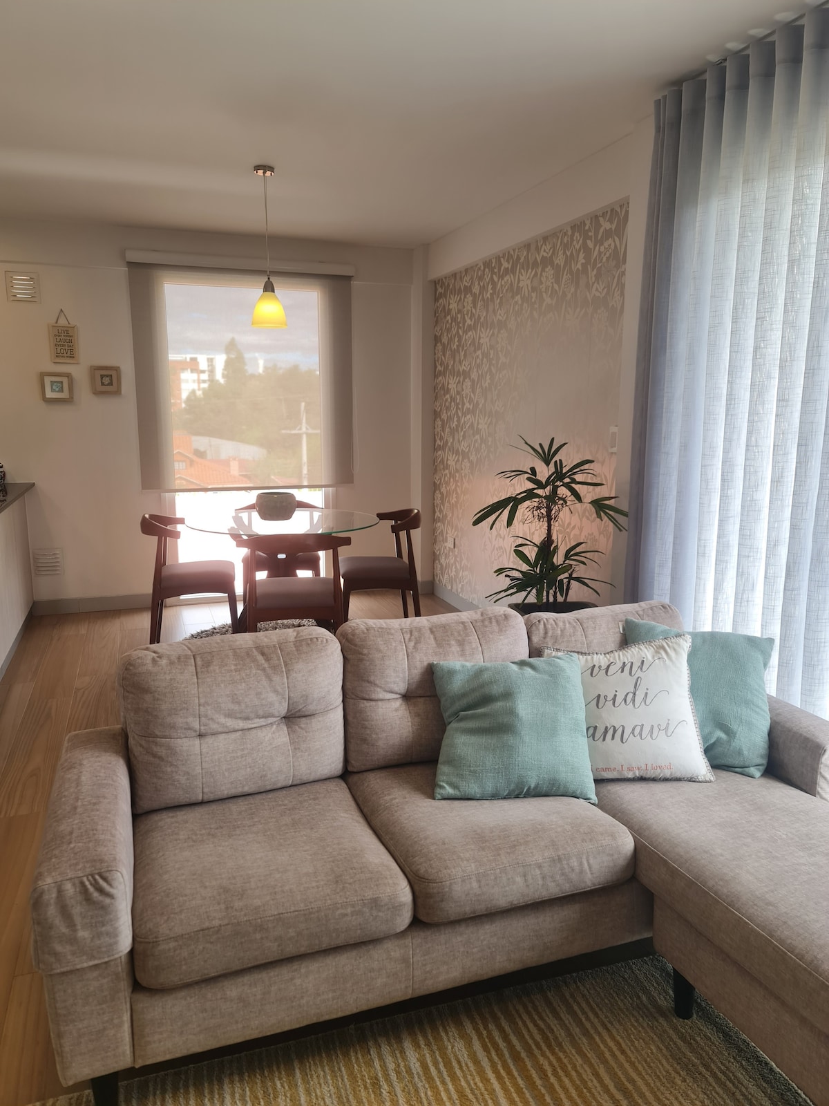 A bright and airy living area features a comfortable sectional sofa adorned with soft cushions. A round dining table with wooden chairs is situated nearby, complemented by a decorative plant. Sheer curtains allow natural light to filter in, enhancing the cheerful ambiance.