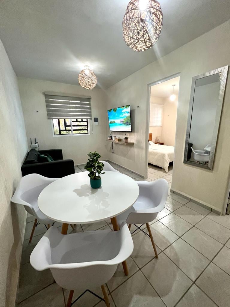 A dining area is displayed featuring a round white table surrounded by four chairs. A small potted plant is centered on the table. A television mounted on the wall displays a serene landscape. Light fixtures with woven designs hang from the ceiling.