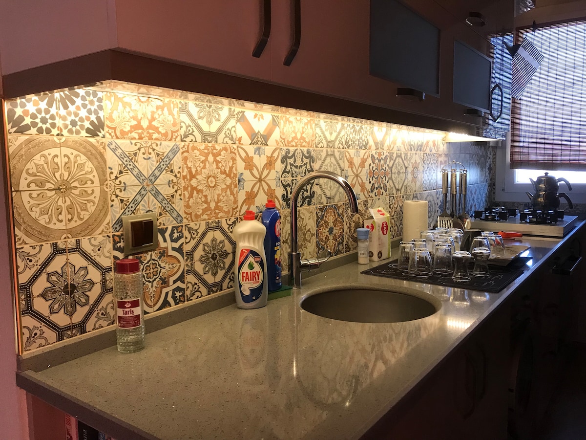 The kitchen displays a well-organized countertop featuring a modern sink, an array of cleaning supplies, and dining utensils. The vibrant tile backsplash adds color, while soft lighting enhances the workspace. Stainless steel appliances are positioned conveniently for easy access.