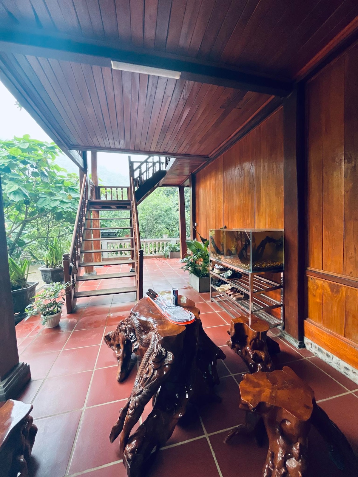 An outdoor terrace features wooden furniture with organic shapes and a staircase leading to the upper level. Lush greenery surrounds the space, creating a serene environment. A large window allows natural light to enhance the warmth of the wooden walls.