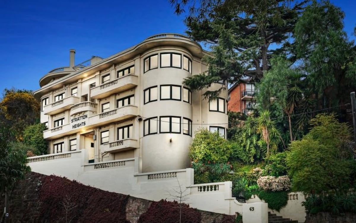 The Stratton Heights apartment building features an elegant façade characterized by rounded architectural elements and an array of large windows. Lush greenery surrounds the structure, providing a harmonious blend with the landscaped garden area in the foreground.