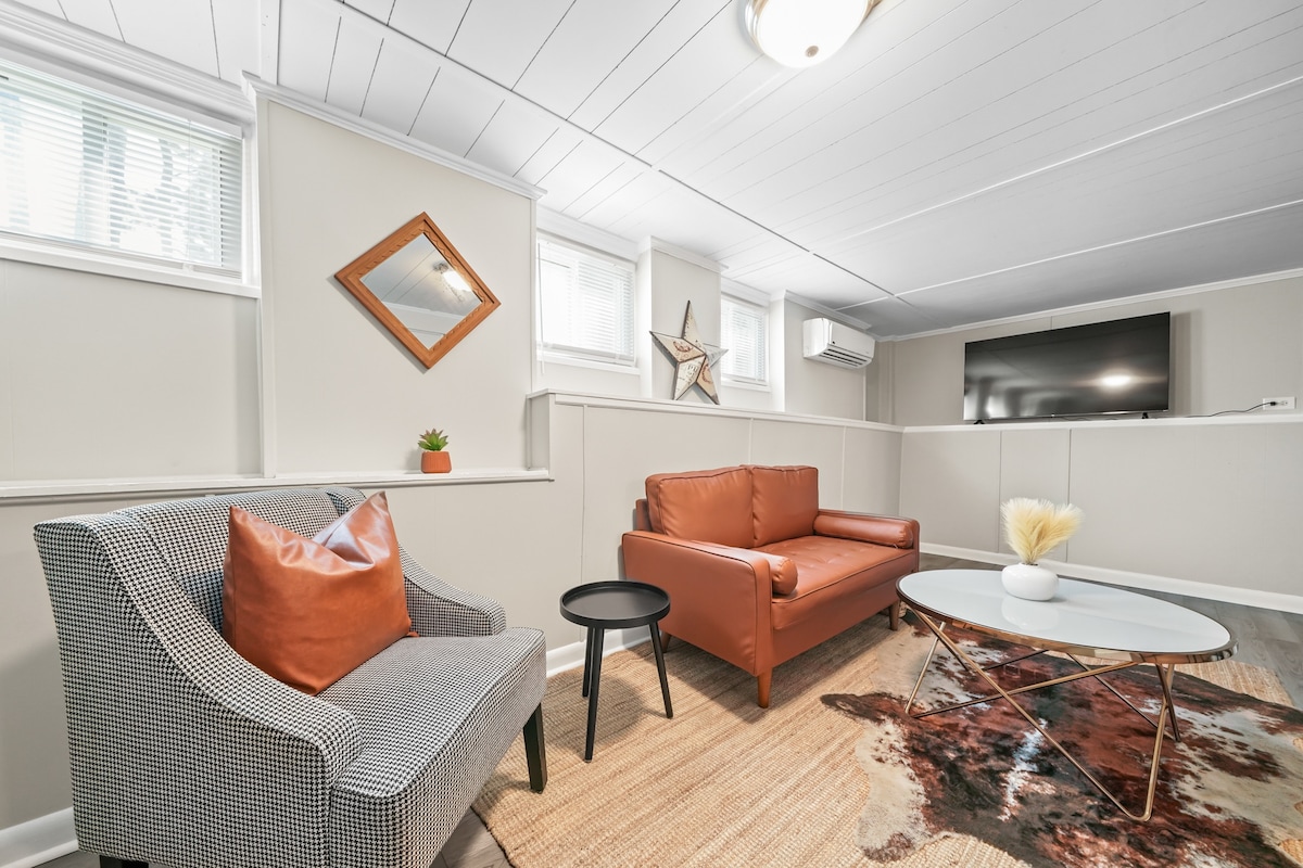 A cozy living area features a brown leather sofa and a patterned armchair with an orange accent pillow. A glass coffee table is centered on a decorative rug, and a wall-mounted TV is visible above a white sideboard. Natural light enters through several windows.