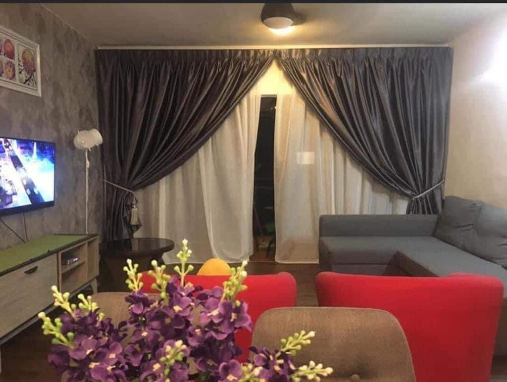 A living area is showcased, featuring a grey sectional sofa and two red chairs. A television is mounted on the wall beside a light wooden media unit. Sheer and heavy curtains frame a doorway, allowing natural light to filter through. A vase with purple flowers adds a touch of color.