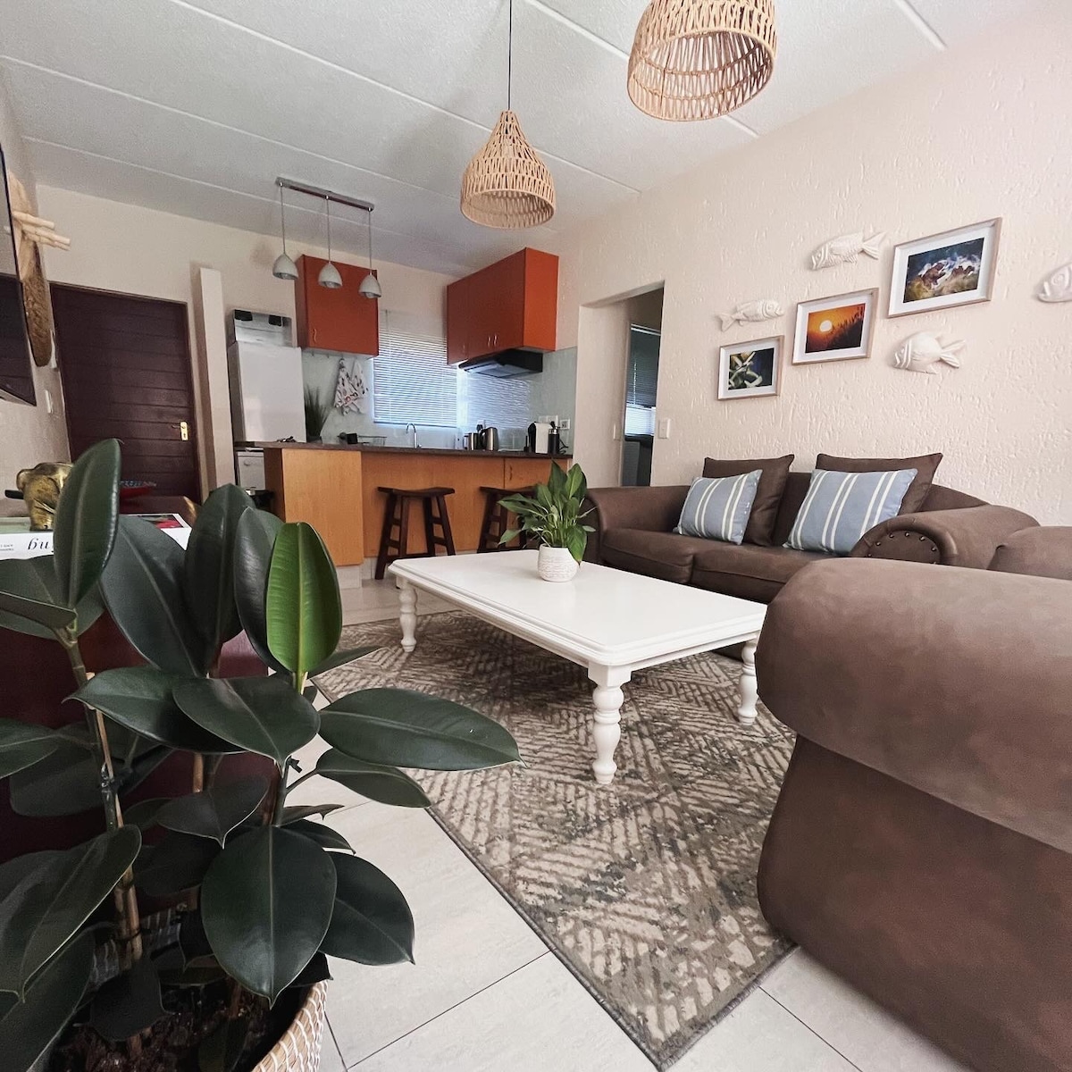 A cozy living area features a comfortable brown sofa with blue accent pillows, positioned near a low, white coffee table. Neutral tones dominate the decor, complemented by a patterned area rug and greenery from nearby potted plants. The kitchen is visible in the background, providing a sense of openness.
