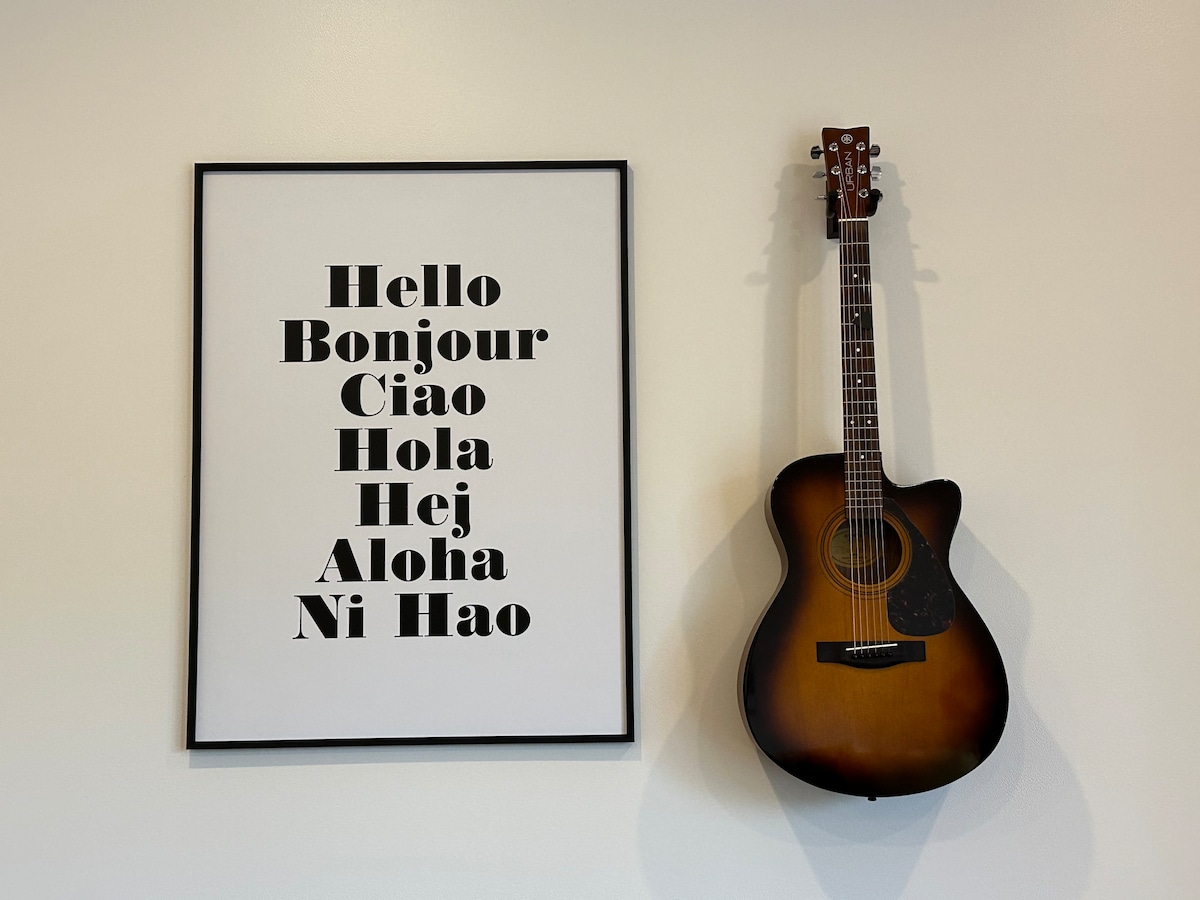 A wall-mounted acoustic guitar is displayed beside a framed print featuring diverse greetings in multiple languages. The print's bold typography contrasts with the soft tones of the guitar, creating an inviting focal point in the space.