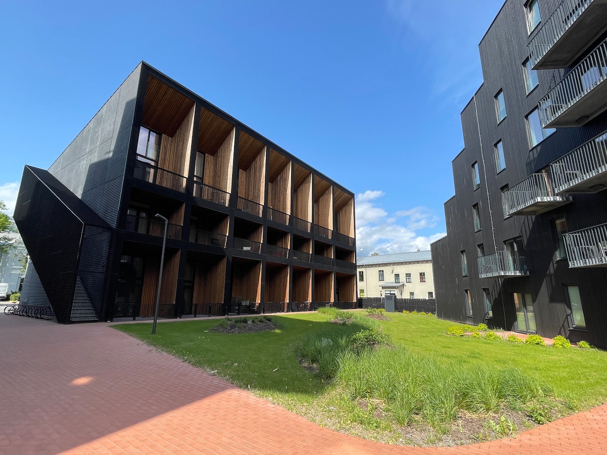 A modern multi-unit building is shown, characterized by its sleek black exterior and wooden accents. The surrounding area features landscaped greenery and paved walkways, with a sunny sky complementing the architectural design.