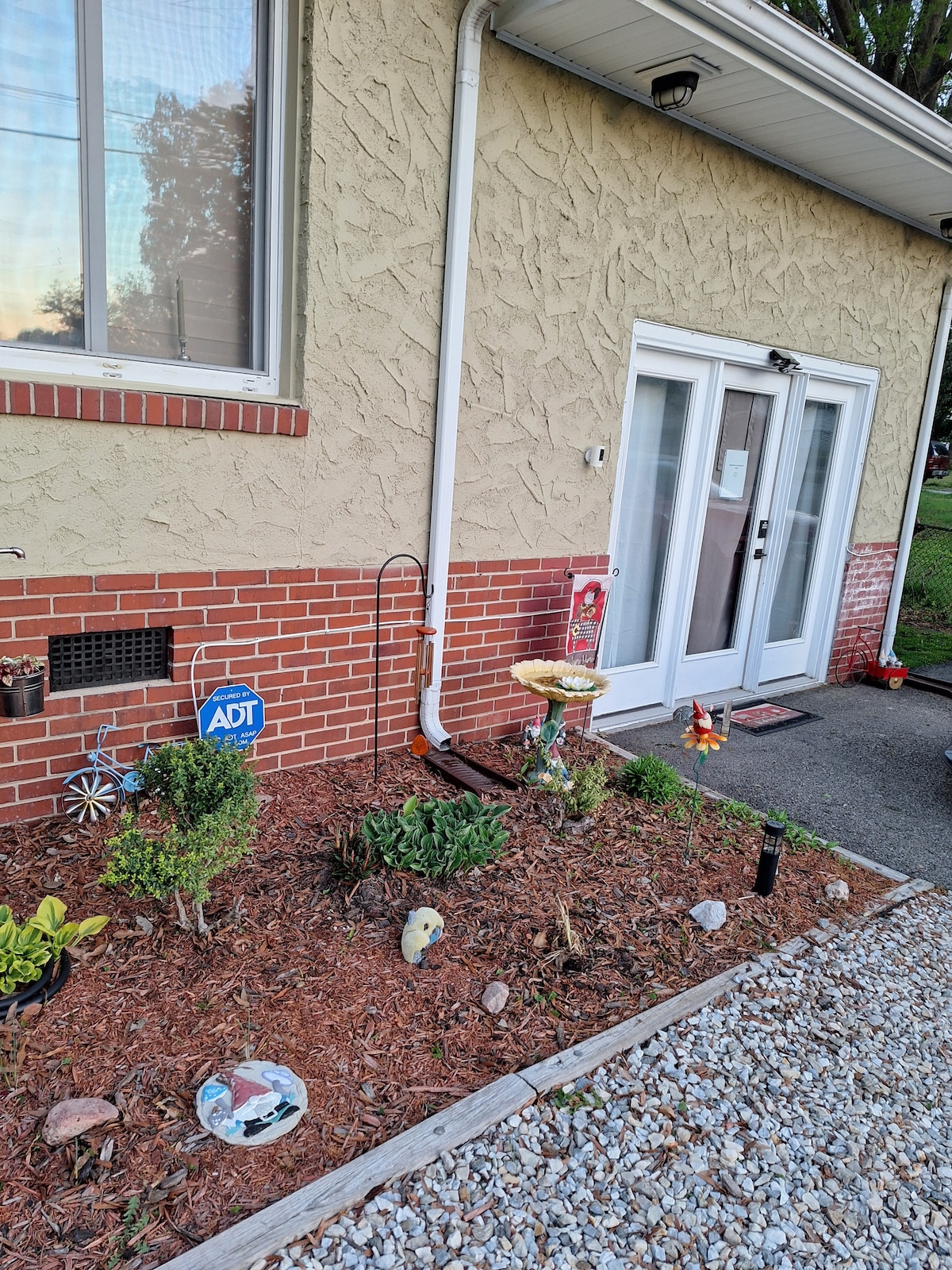 The entrance features a well-kept garden area with decorative stones and plants. Two glass doors provide access to the interior, framed by a light-colored exterior wall. A security sign is visible, along with additional decorative elements enhancing the entrance's inviting look.