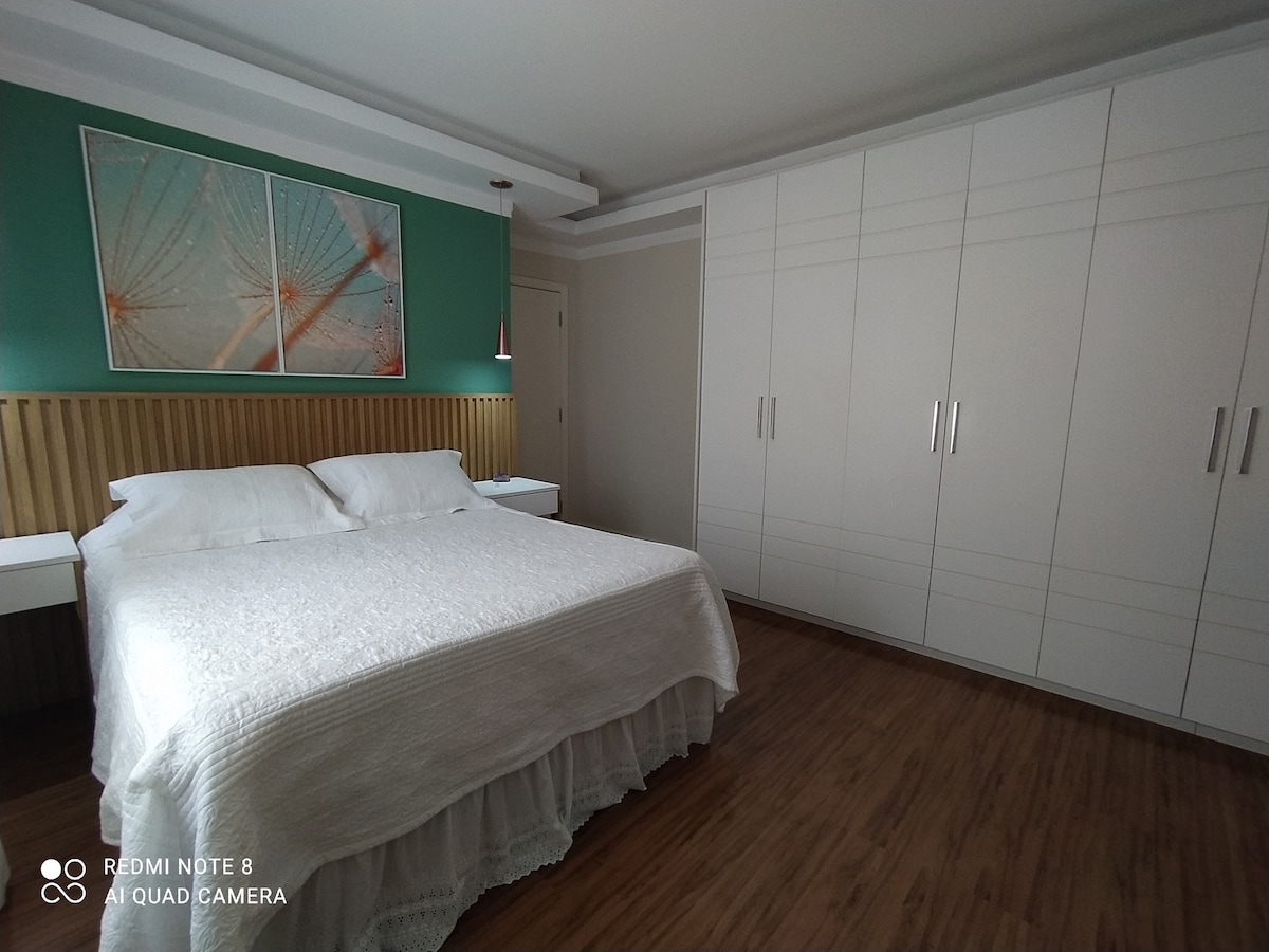 A well-arranged bedroom is presented, featuring a spacious bed with a light-colored duvet. Two bedside tables with lamps flank the bed. A large wardrobe offers ample storage, while a piece of artwork adorns the green accent wall above the headboard.