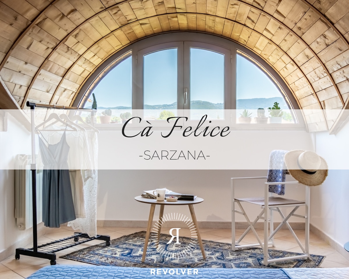 A spacious bedroom features a striking wooden barrel ceiling and a large window with a view of the surrounding hills. A clothing rack displays garments, while a small chair and table are positioned on a textured rug, providing a cozy and functional space.