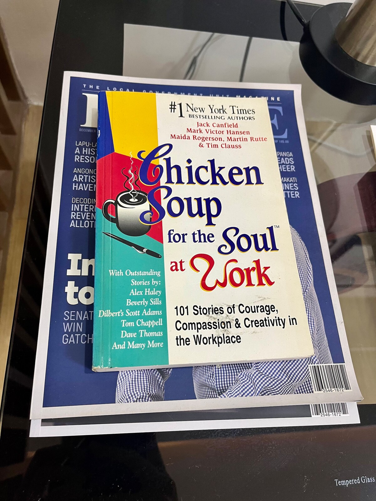 A stack of magazines is arranged on a glass surface. The top magazine features a bright cover of 'Chicken Soup for the Soul at Work,' showcasing colorful graphics and bold text. The layout is clean, suggesting a dedicated workspace.