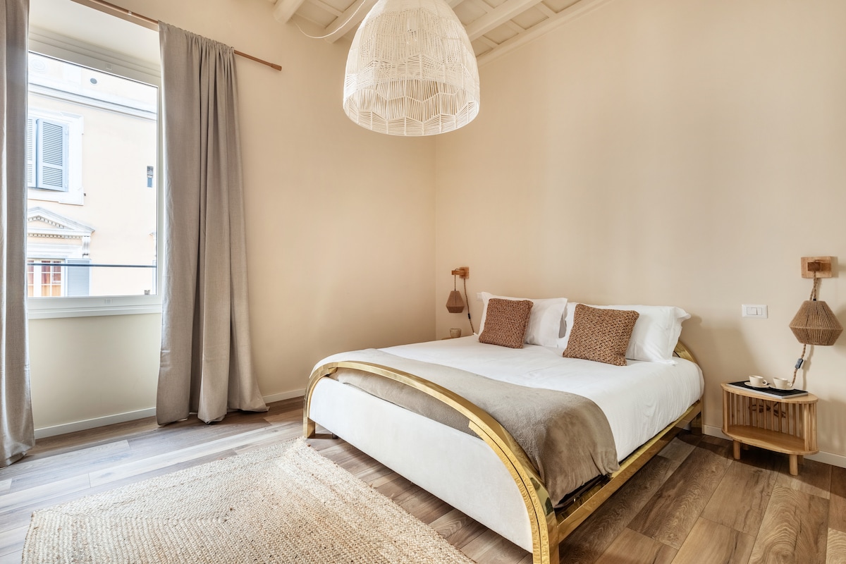 A spacious bedroom features a light wood bed with neutral bedding. Two decorative pillows add a subtle touch. Natural light enters through a window adorned with soft curtains. A woven area rug and minimalist nightstands complete the serene atmosphere.