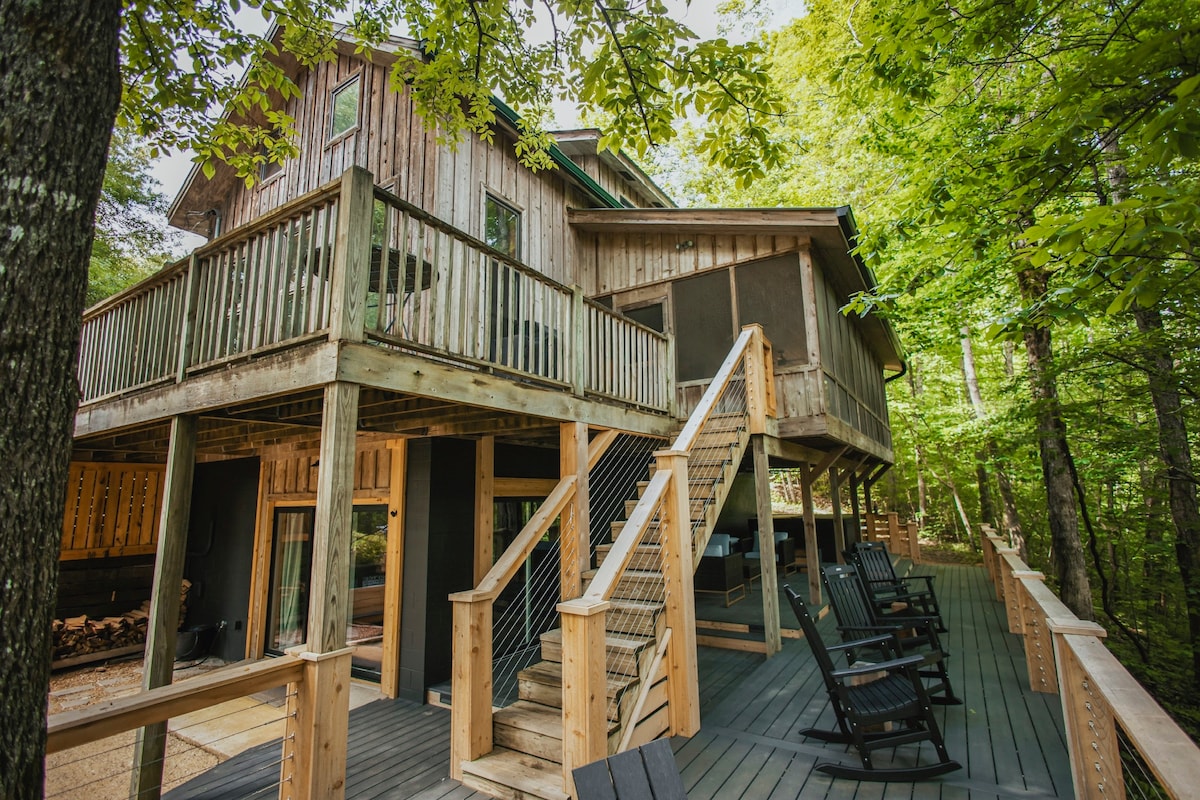 A spacious cabin is nestled among lush trees, featuring multiple levels and a large outdoor deck. Sturdy wooden stairs lead to the upper level, where a screened porch offers relaxing seating. The inviting, natural surroundings enhance the peaceful atmosphere of the retreat.