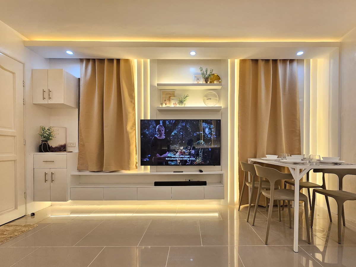 A modern living area is showcased with a large flat-screen television mounted against a white wall, illuminated by soft lighting. Light-colored curtains frame the television, and a dining table with chairs is positioned nearby, creating a cozy atmosphere for gatherings.