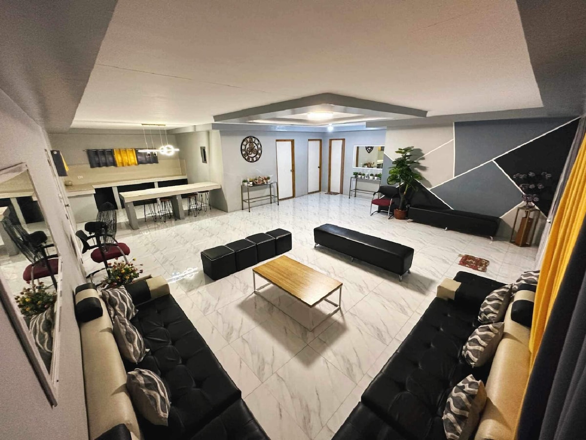 A spacious living area features sleek leather seating arranged around a rectangular coffee table. Light-colored walls brighten the room, and large windows allow natural light to fill the space. A dining area with modern bar stools is visible in the background, enhancing the open layout.