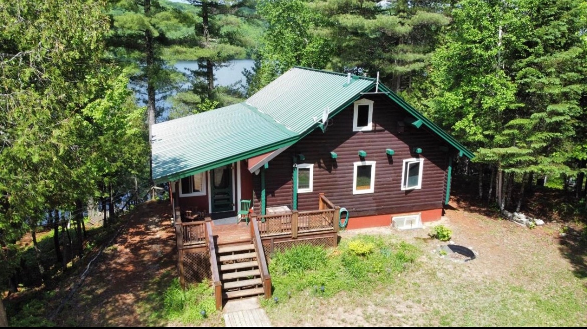 A rustic cabin is showcased, surrounded by a lush forest with a visible lake in the background. The structure features a sloped green roof and wooden deck steps leading to the entrance. Large windows invite natural light into the space, blending the home harmoniously with nature.