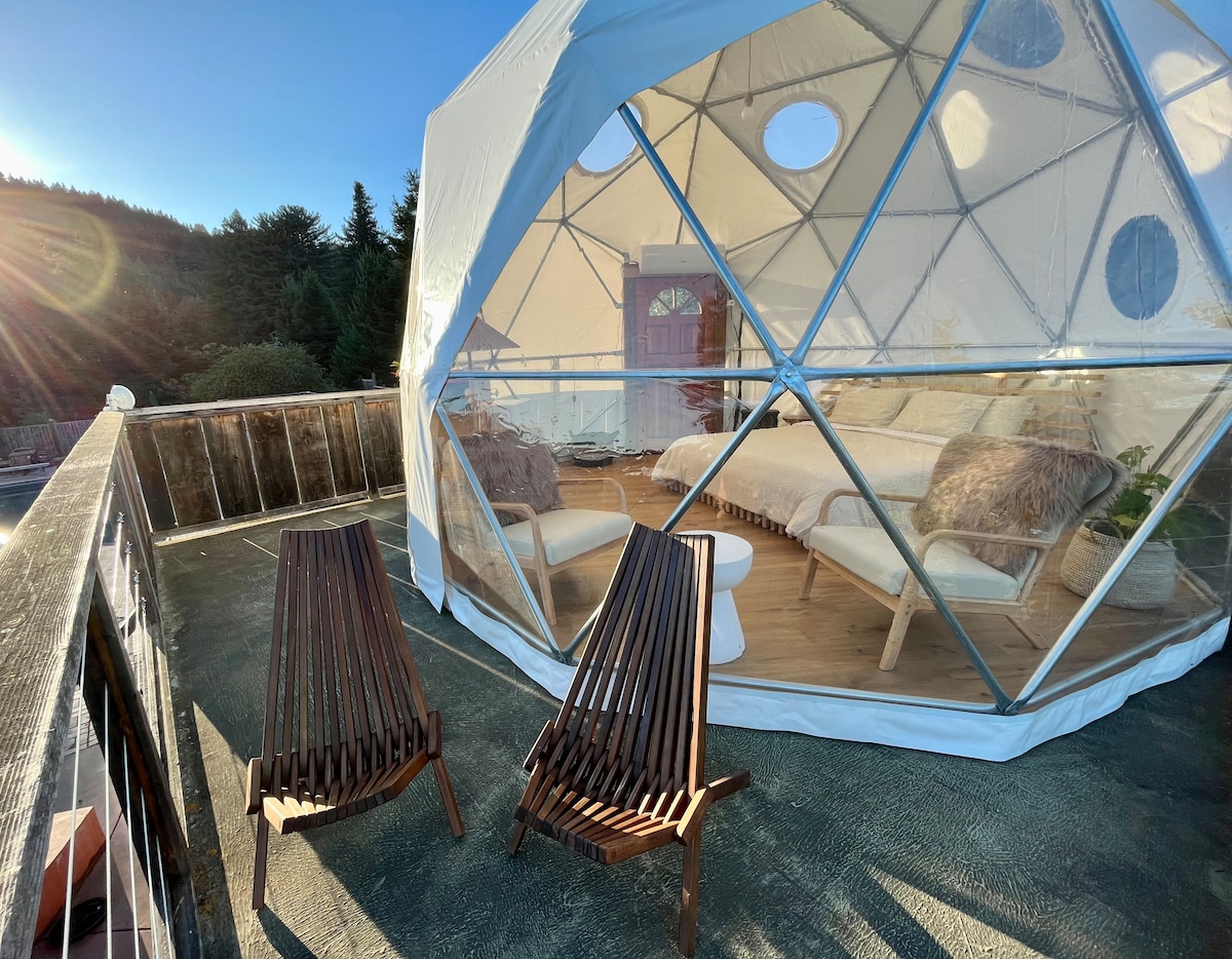 An outdoor deck extends from the dome, featuring two slatted wooden lounge chairs. The dome's transparent structure allows for abundant natural light and provides views of the surrounding hills and trees. A cozy sleeping area is visible inside, inviting relaxation.